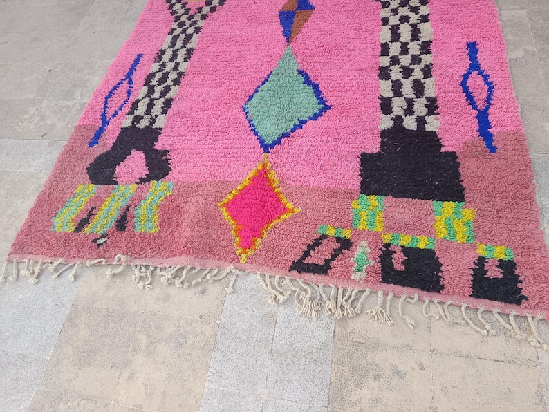 May include: A pink Moroccan rug with geometric designs. The rug features a central pink field with black, blue, yellow, and green geometric shapes. The rug has a fringe along the bottom edge.