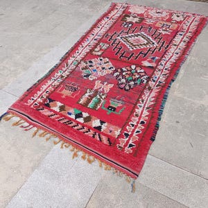 May include: A rectangular red rug with intricate geometric patterns and colorful accents. The rug features a central diamond design, bordered by white and black patterns, and finished with tassels. The rug's dimensions are not visible.