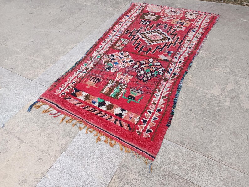 May include: A rectangular red rug with intricate geometric patterns and colorful accents. The rug features a central diamond design, bordered by white and black patterns, and finished with tassels. The rug's dimensions are not visible.