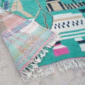 May include: A turquoise and beige geometric patterned rug with a rolled-up section revealing a striped design. The rug features black, white, and pink accents, with a fringe along the edge. The rug's design includes shapes and lines.