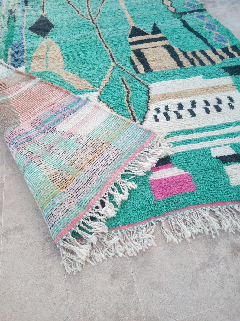May include: A turquoise and beige geometric patterned rug with a rolled-up section revealing a striped design. The rug features black, white, and pink accents, with a fringe along the edge. The rug's design includes shapes and lines.