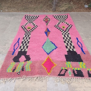 May include: A vibrant pink Moroccan rug with geometric patterns. The rug features black and white checkered designs, diamonds, and other shapes in blue, green, yellow, and pink. The rug has a fringed edge.