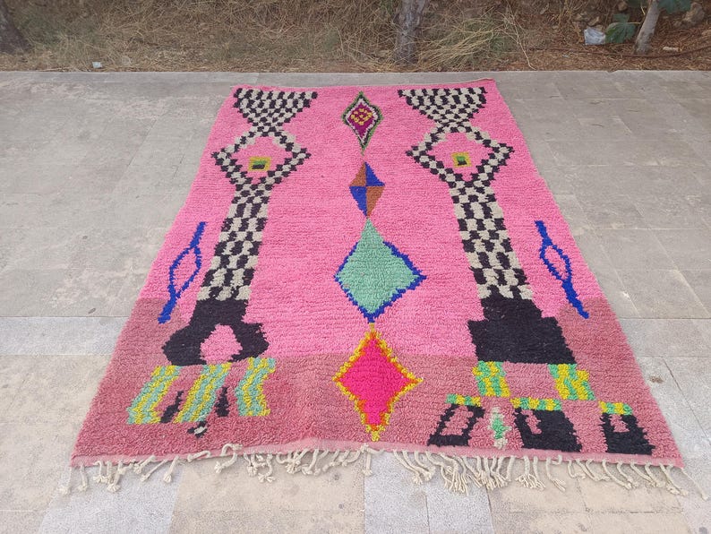 May include: A vibrant pink Moroccan rug with geometric patterns. The rug features black and white checkered designs, diamonds, and other shapes in blue, green, yellow, and pink. The rug has a fringed edge.