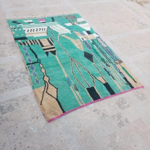 May include: A rectangular rug with a teal background and an abstract design. The rug features geometric shapes in black, beige, and blue, with a pink border. The rug has a fringe on one end and is placed on a concrete surface.