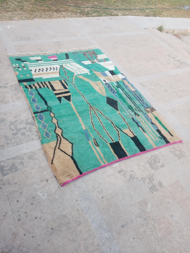 May include: A rectangular rug with a teal background and an abstract design. The rug features geometric shapes in black, beige, and blue, with a pink border. The rug has a fringe on one end and is placed on a concrete surface.