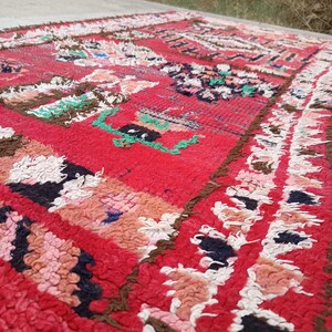 May include: A vibrant red rug with a textured, woven design. The rug features geometric patterns in shades of pink, white, brown, green, and black. The rug's edges have a fringed border.