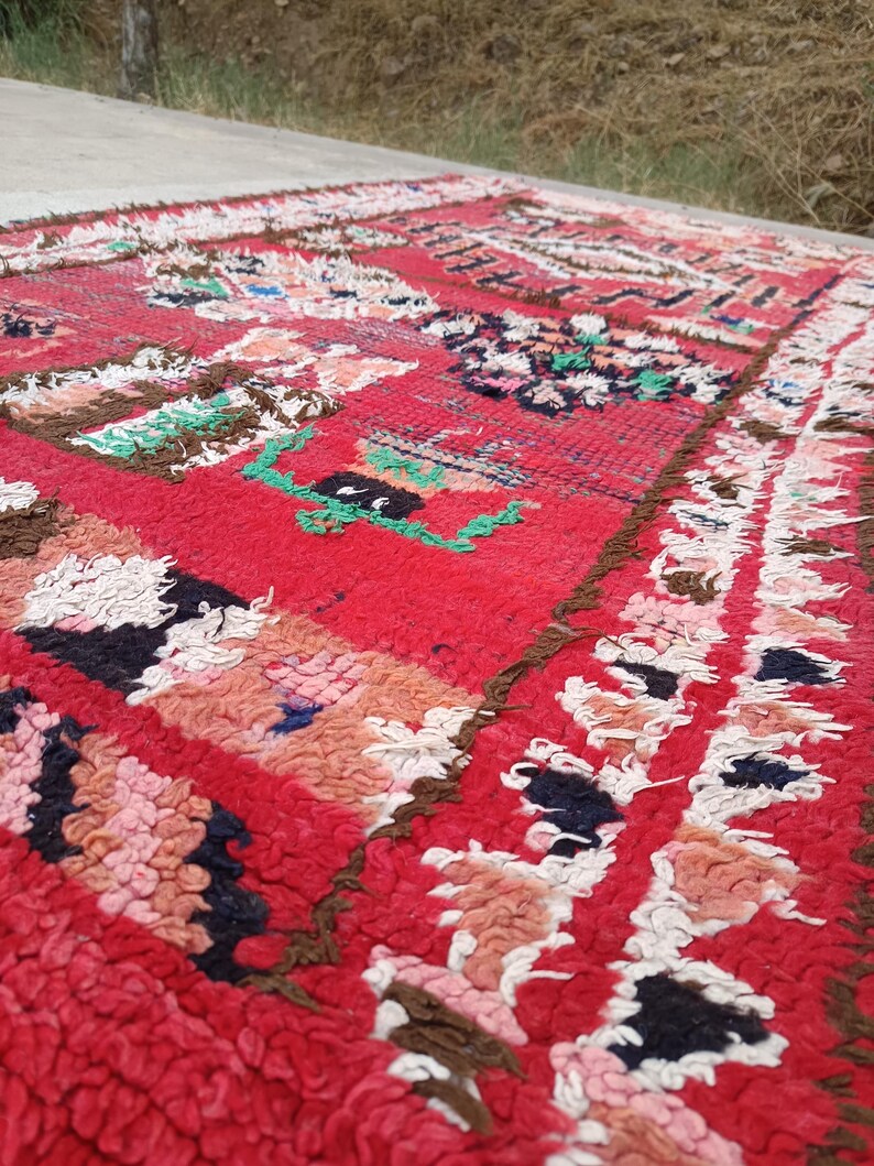 May include: A vibrant red rug with a textured, woven design. The rug features geometric patterns in shades of pink, white, brown, green, and black. The rug's edges have a fringed border.