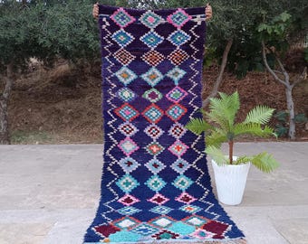 May include: A long, rectangular rug with a dark blue background and a repeating diamond pattern in various colors. The diamonds are filled with shades of pink, green, orange, and white. The rug has a fringed edge.