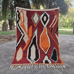 May include: A rectangular, hand-woven rug with a deep red background and geometric patterns in black, white, and orange. The rug features diamond and teardrop shapes, with a fringe along the bottom edge. The rug is displayed outdoors.