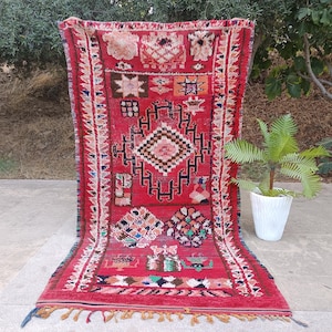 May include: A vibrant red Moroccan rug with intricate geometric patterns and a fringed edge. The rug features a central diamond design and colorful accents in pink, blue, and green. The rug is displayed outdoors, showcasing its texture and design.