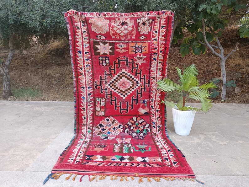 May include: A vibrant red Moroccan rug with intricate geometric patterns and a fringed edge. The rug features a central diamond design and colorful accents in pink, blue, and green. The rug is displayed outdoors, showcasing its texture and design.