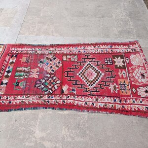May include: A rectangular red rug with geometric patterns and fringe. The rug features a central diamond design with black and white accents, surrounded by smaller colorful shapes and a white border. The rug is placed on a gray surface.