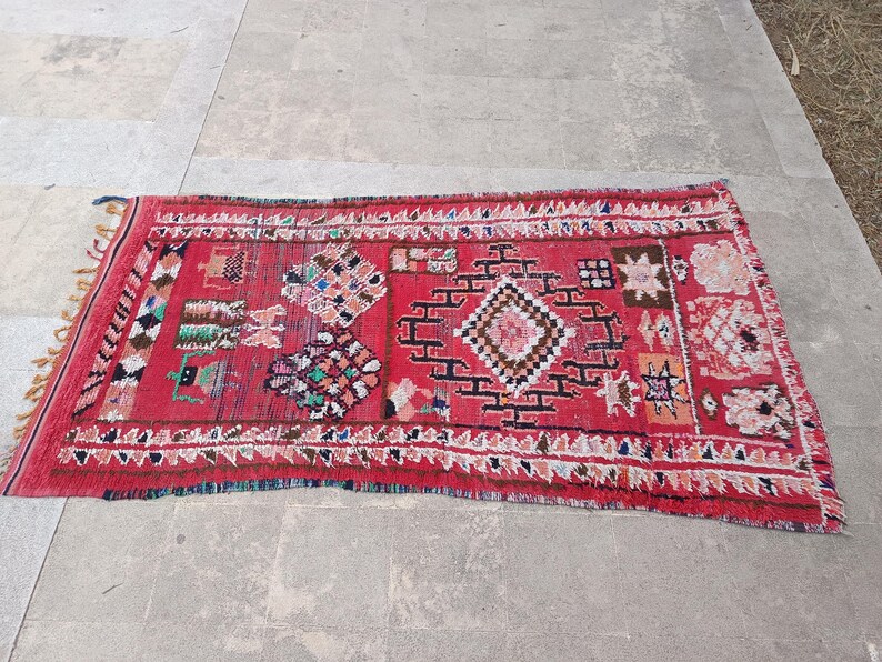 May include: A rectangular red rug with geometric patterns and fringe. The rug features a central diamond design with black and white accents, surrounded by smaller colorful shapes and a white border. The rug is placed on a gray surface.