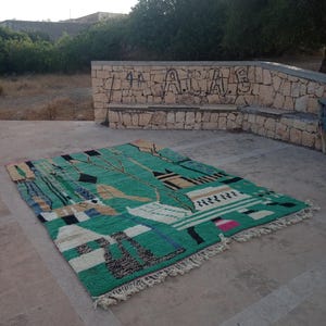 May include: A rectangular, hand-woven rug with a vibrant green background and a geometric design. The rug features black, white, tan, and blue shapes, with a fringed edge. The rug is displayed outdoors on a concrete surface.