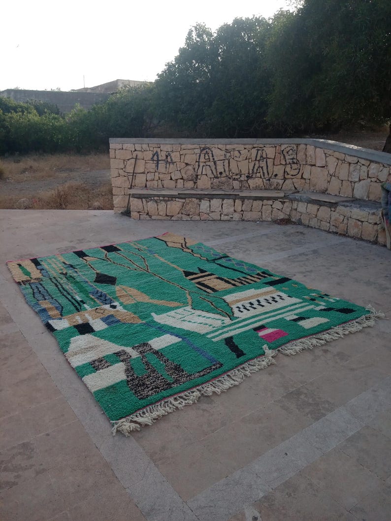 May include: A rectangular, hand-woven rug with a vibrant green background and a geometric design. The rug features black, white, tan, and blue shapes, with a fringed edge. The rug is displayed outdoors on a concrete surface.