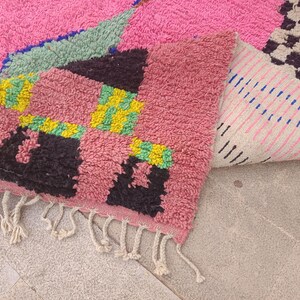 May include: A vibrant, hand-woven rug with a pink, green, black, and yellow geometric design. The rug features a soft, textured surface and a fringed edge. The folded section reveals a striped pattern on the underside.