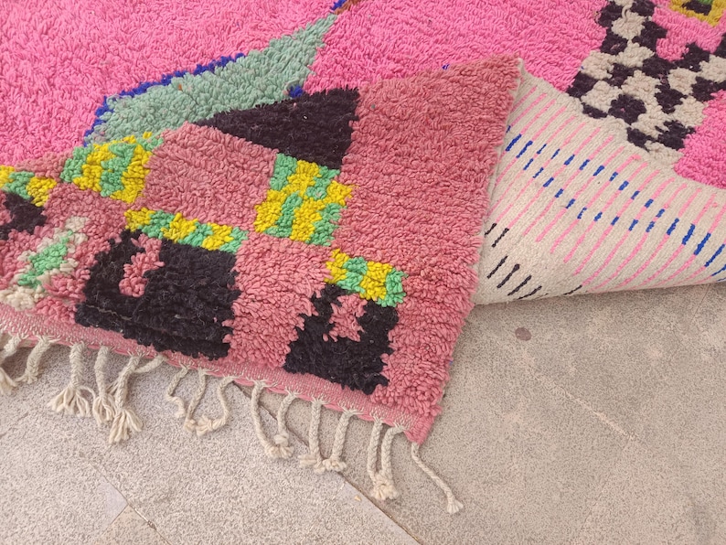 May include: A vibrant, hand-woven rug with a pink, green, black, and yellow geometric design. The rug features a soft, textured surface and a fringed edge. The folded section reveals a striped pattern on the underside.