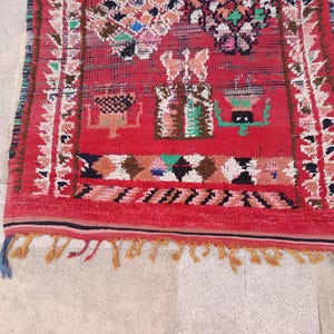 May include: A vibrant red rug with intricate geometric patterns and a fringed edge. The rug features a central design with colorful motifs in green, white, and brown. The border is adorned with a repeating pattern in white and brown. The rug's design is a blend of traditional and contemporary styles.