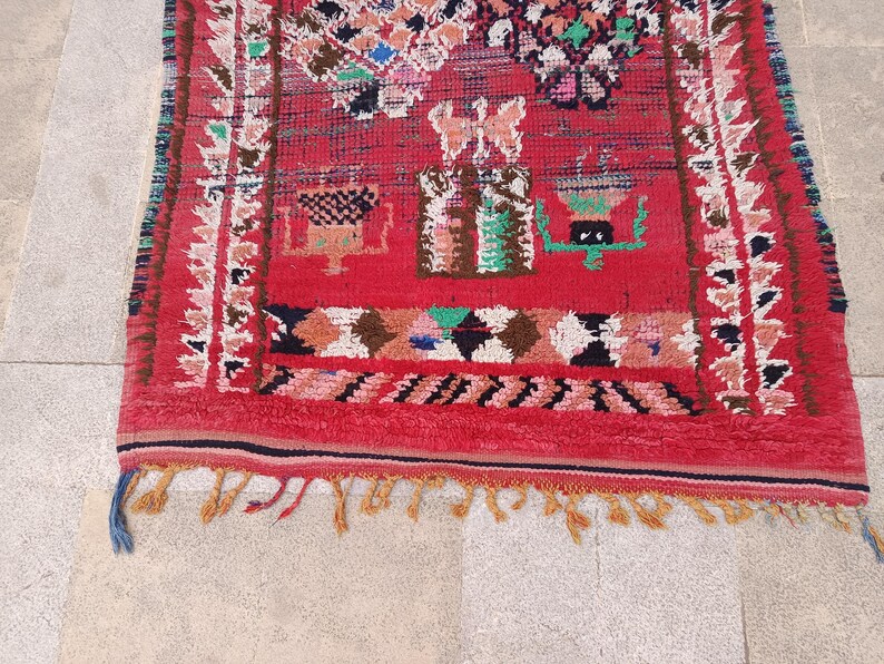 May include: A vibrant red rug with intricate geometric patterns and a fringed edge. The rug features a central design with colorful motifs in green, white, and brown. The border is adorned with a repeating pattern in white and brown. The rug's design is a blend of traditional and contemporary styles.