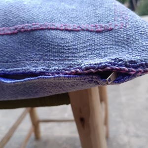 May include: Close-up of a blue woven pillow with a pink stitched design. The pillow has a zipper closure and is resting on a wooden stool. The fabric has a textured appearance.