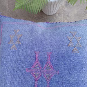 May include: A square, blue woven floor cushion with geometric patterns in pink and tan. The cushion has a textured surface and is likely made of natural fibers. The design features symmetrical shapes, and the edges are finished with a pink trim.