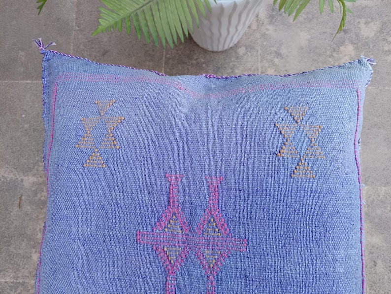 May include: A square, blue woven floor cushion with geometric patterns in pink and tan. The cushion has a textured surface and is likely made of natural fibers. The design features symmetrical shapes, and the edges are finished with a pink trim.