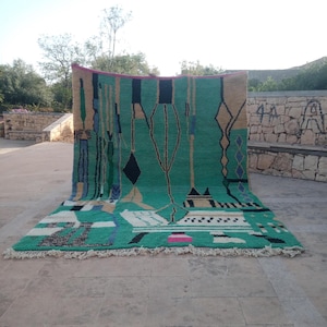 May include: A large, rectangular rug with a vibrant green central field. The rug features geometric patterns in black, beige, blue, and white, with a fringe along the bottom edge. The design includes diamond and tree-like motifs.