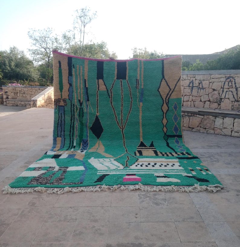 May include: A large, rectangular rug with a vibrant green central field. The rug features geometric patterns in black, beige, blue, and white, with a fringe along the bottom edge. The design includes diamond and tree-like motifs.