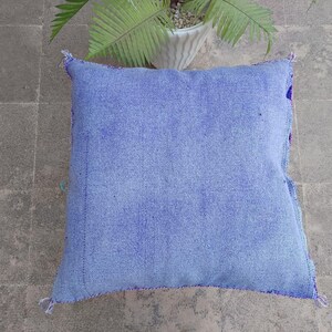 May include: A square, light blue cushion with a textured fabric. The cushion has a stitched border in a darker blue and purple. The background is a gray tiled surface with a potted plant.