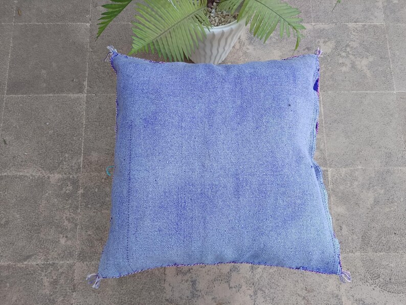 May include: A square, light blue cushion with a textured fabric. The cushion has a stitched border in a darker blue and purple. The background is a gray tiled surface with a potted plant.