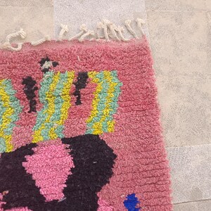 May include: A close-up of a pink rug with a geometric design. The rug features a pattern of black, yellow, and blue shapes against a pink background. The rug has a textured, woven appearance and a fringe along the top edge.