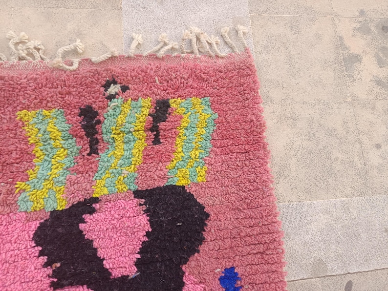 May include: A close-up of a pink rug with a geometric design. The rug features a pattern of black, yellow, and blue shapes against a pink background. The rug has a textured, woven appearance and a fringe along the top edge.