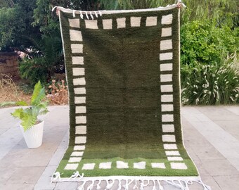 May include: A rectangular green rug with a white border featuring a series of white rectangular blocks. The rug has a fringe at the bottom and is displayed outdoors. The rug's design is simple and geometric.