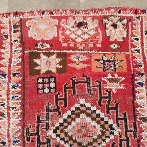 May include: A vibrant, hand-woven rug with a red background and intricate geometric patterns in shades of brown, white, and orange. The rug features a fringed border and a textured surface, showcasing traditional craftsmanship.