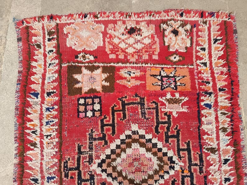 May include: A vibrant, hand-woven rug with a red background and intricate geometric patterns in shades of brown, white, and orange. The rug features a fringed border and a textured surface, showcasing traditional craftsmanship.