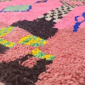 May include: Close-up of a pink, textured rug with geometric patterns. The rug features black, yellow, green, blue, and white shapes. The rug has a thick, plush pile, suggesting a soft and comfortable texture. The design is abstract and colorful.