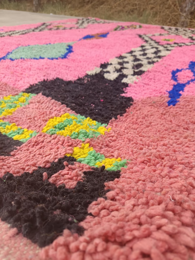 May include: Close-up of a pink, textured rug with geometric patterns. The rug features black, yellow, green, blue, and white shapes. The rug has a thick, plush pile, suggesting a soft and comfortable texture. The design is abstract and colorful.
