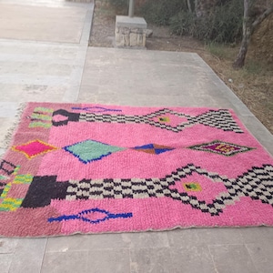 May include: A vibrant pink rug with geometric patterns. The rug features diamond and rectangular shapes in black, white, blue, green, and yellow. The rug has a fringed edge and is placed on a concrete surface.