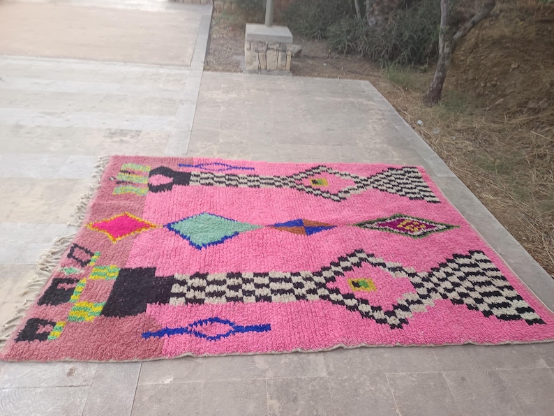 May include: A vibrant pink rug with geometric patterns. The rug features diamond and rectangular shapes in black, white, blue, green, and yellow. The rug has a fringed edge and is placed on a concrete surface.