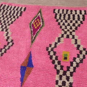 May include: A pink rug with geometric patterns. The rug features repeating black and white checkered designs, diamond shapes, and colorful accents in green, blue, and yellow. The rug's texture appears soft and plush.