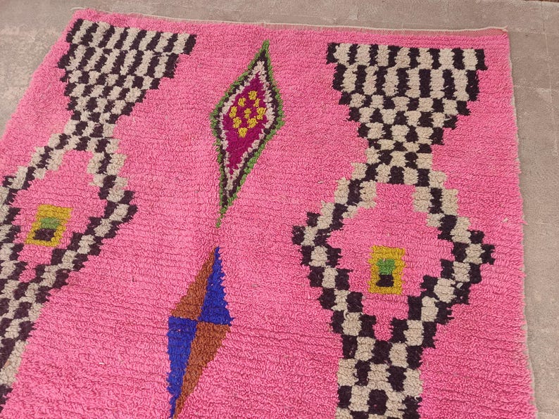 May include: A pink rug with geometric patterns. The rug features repeating black and white checkered designs, diamond shapes, and colorful accents in green, blue, and yellow. The rug's texture appears soft and plush.