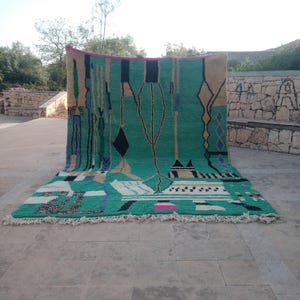 May include: A large, rectangular rug with a vibrant green central field. The rug features geometric patterns in black, beige, blue, and pink. The rug has a fringe along the bottom edge and is displayed outdoors.