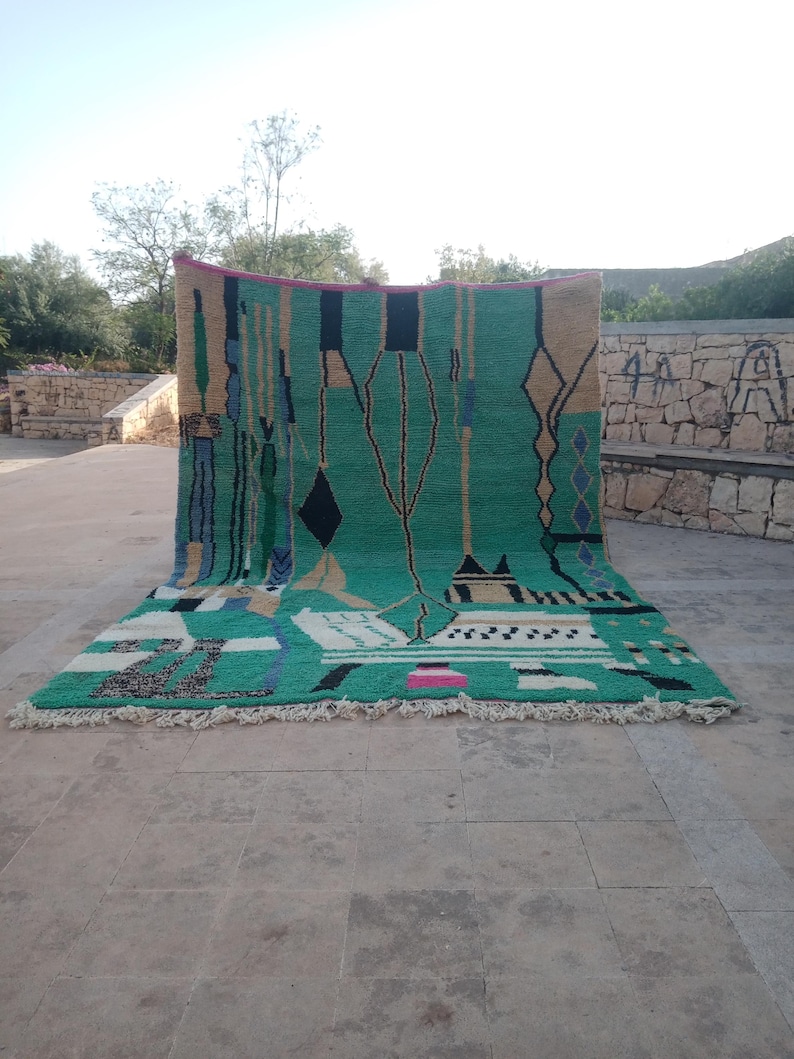 May include: A large, rectangular rug with a vibrant green central field. The rug features geometric patterns in black, beige, blue, and pink. The rug has a fringe along the bottom edge and is displayed outdoors.