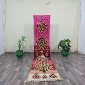May include: A colourful pink and red runner rug with diamond patterns. The rug has green, orange, and white details, and a fringed edge. It's displayed on a wooden floor, with potted plants on either side. The rug's length is approximately 200 cm.