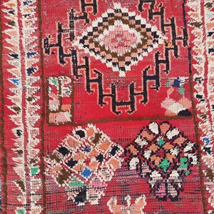 May include: A vibrant red rug with intricate geometric patterns in black, white, brown, and various other colors. The rug features a central diamond-shaped design and floral motifs, with a fringed border.