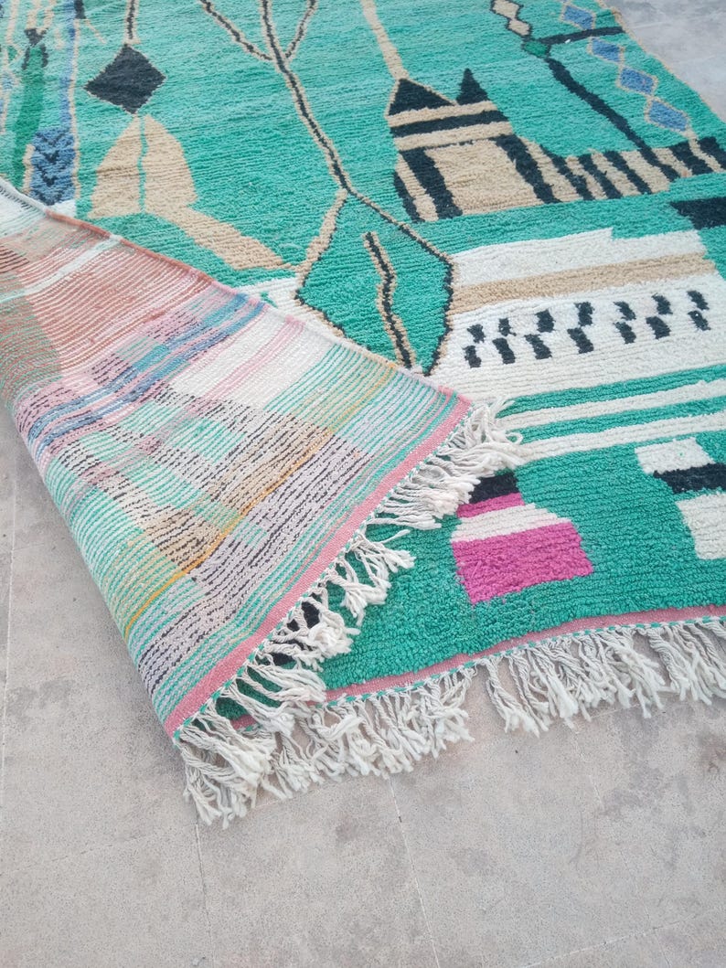 May include: A turquoise and beige geometric patterned rug with a rolled-up section revealing a striped design. The rug features black, white, and pink accents, with a fringe along the edge. The rug's design includes shapes and lines.