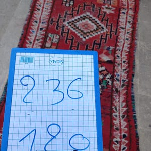 May include: A rectangular, red rug with a geometric pattern and fringe. The rug features a central diamond design and various other shapes in black, white, and orange. A blue and white grid board with the numbers 236 and 180 is in the foreground.