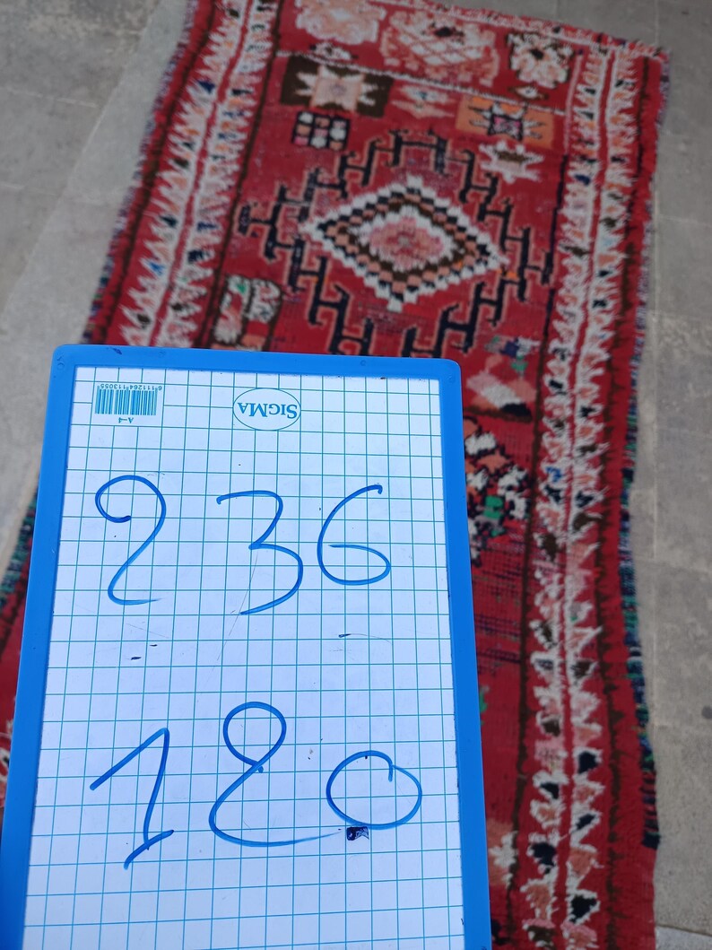 May include: A rectangular, red rug with a geometric pattern and fringe. The rug features a central diamond design and various other shapes in black, white, and orange. A blue and white grid board with the numbers 236 and 180 is in the foreground.