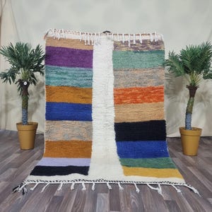 Könnte beinhalten: Ein rechteckiger, mehrfarbiger Teppich mit Streifenmuster. Der Teppich hat horizontale Bänder in verschiedenen Farben, darunter Lila, Blau, Orange und Schwarz, die durch einen vertikalen weißen Streifen getrennt sind. Der Teppich hat einen Fransenrand.