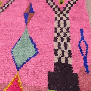 May include: A vibrant pink rug with geometric patterns. The design includes diamond and rectangular shapes in blue, green, brown, and black. The rug has a textured surface and a colorful, eye-catching design.